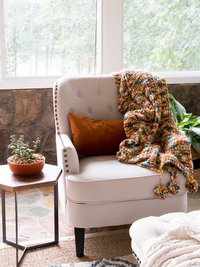 Cream color accent chair with throw blanket with autumn colors and rust lumbar pillow.