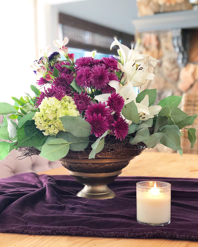 Fall floral arrangement with plum colors sitting on a plum table runner with candle.