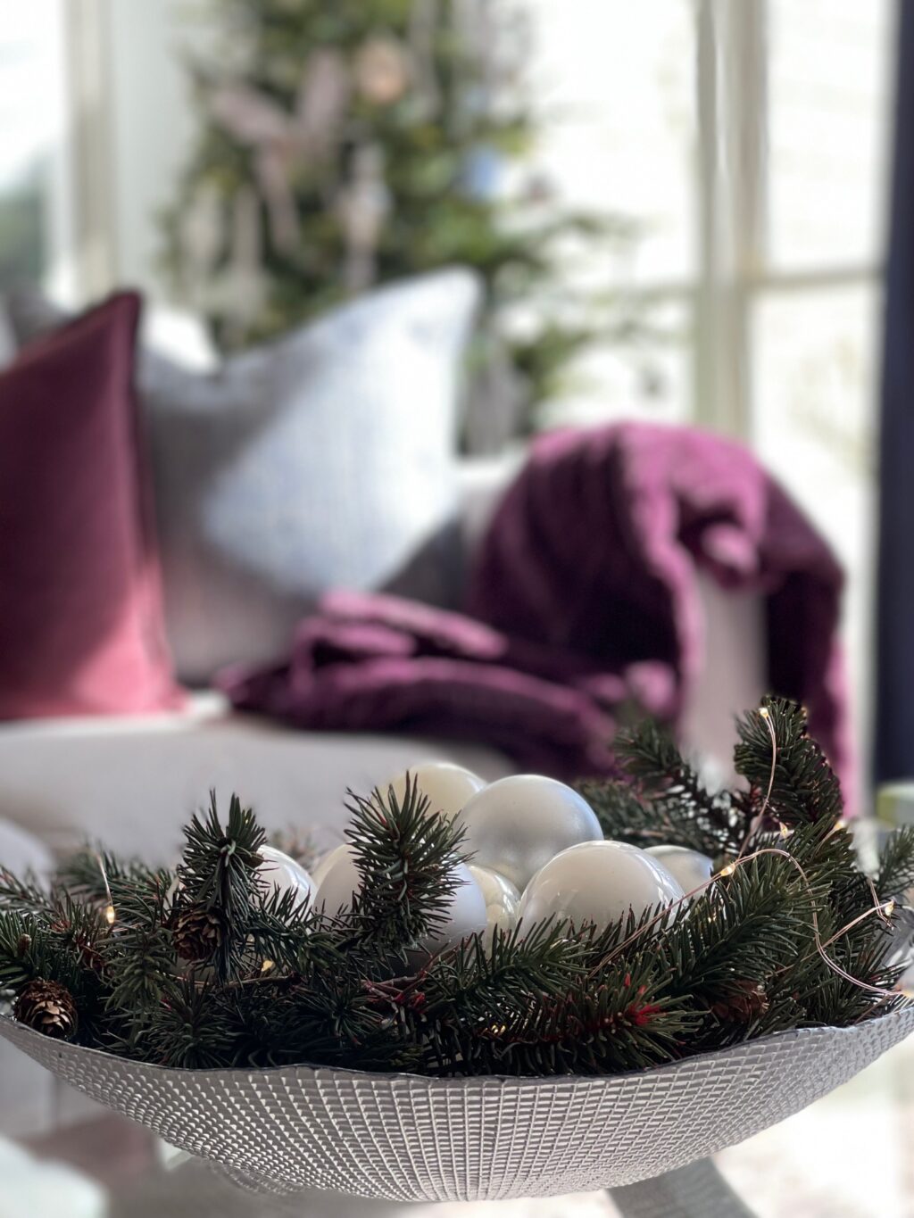 bowl filled with ever green and ornaments with decorated tree in background.
